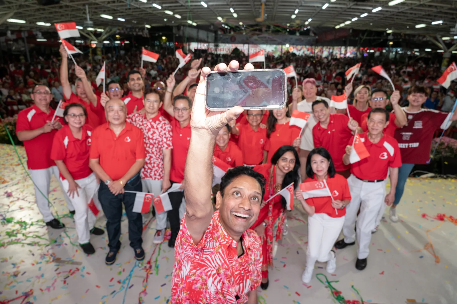 High-energy crowd celebration with flags, confetti and a well-timed selfie foreground subject — genuine event atmosphere and depth