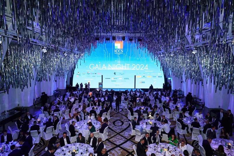 Wide gala ballroom under hanging silver streamers with a lit stage screen above full banquet tables