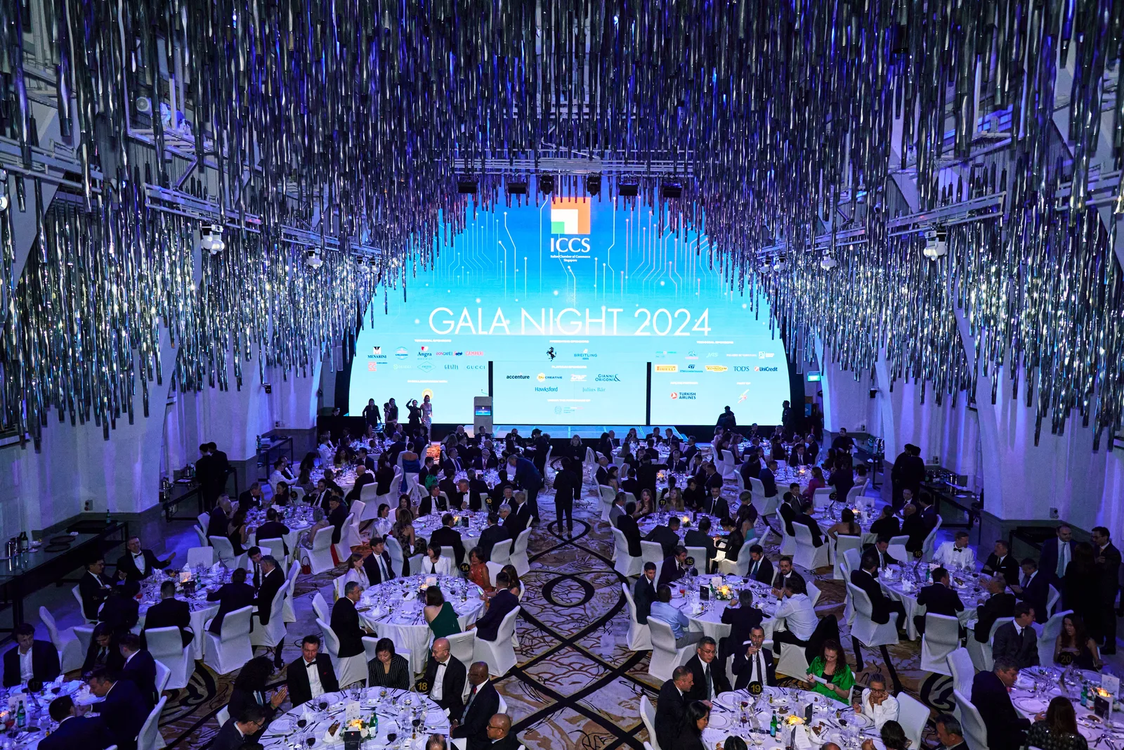 Wide gala ballroom under a ceiling of hanging silver streamers, 'GALA NIGHT 2024' screen glowing over full banquet tables in purple/blue was