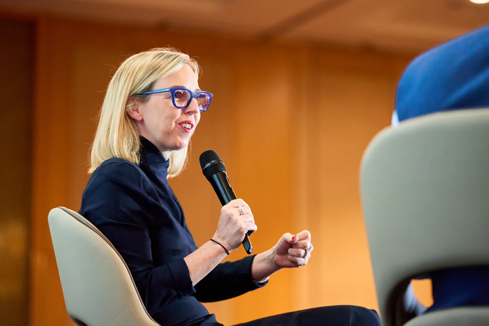 Seated panellist mid-sentence with mic, expressive hands, clean uncluttered background, crisp light
