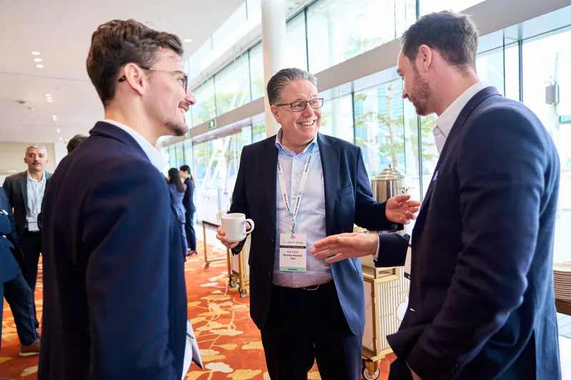 Three delegates in conversation with lanyards and coffee in a bright glass atrium between sessions