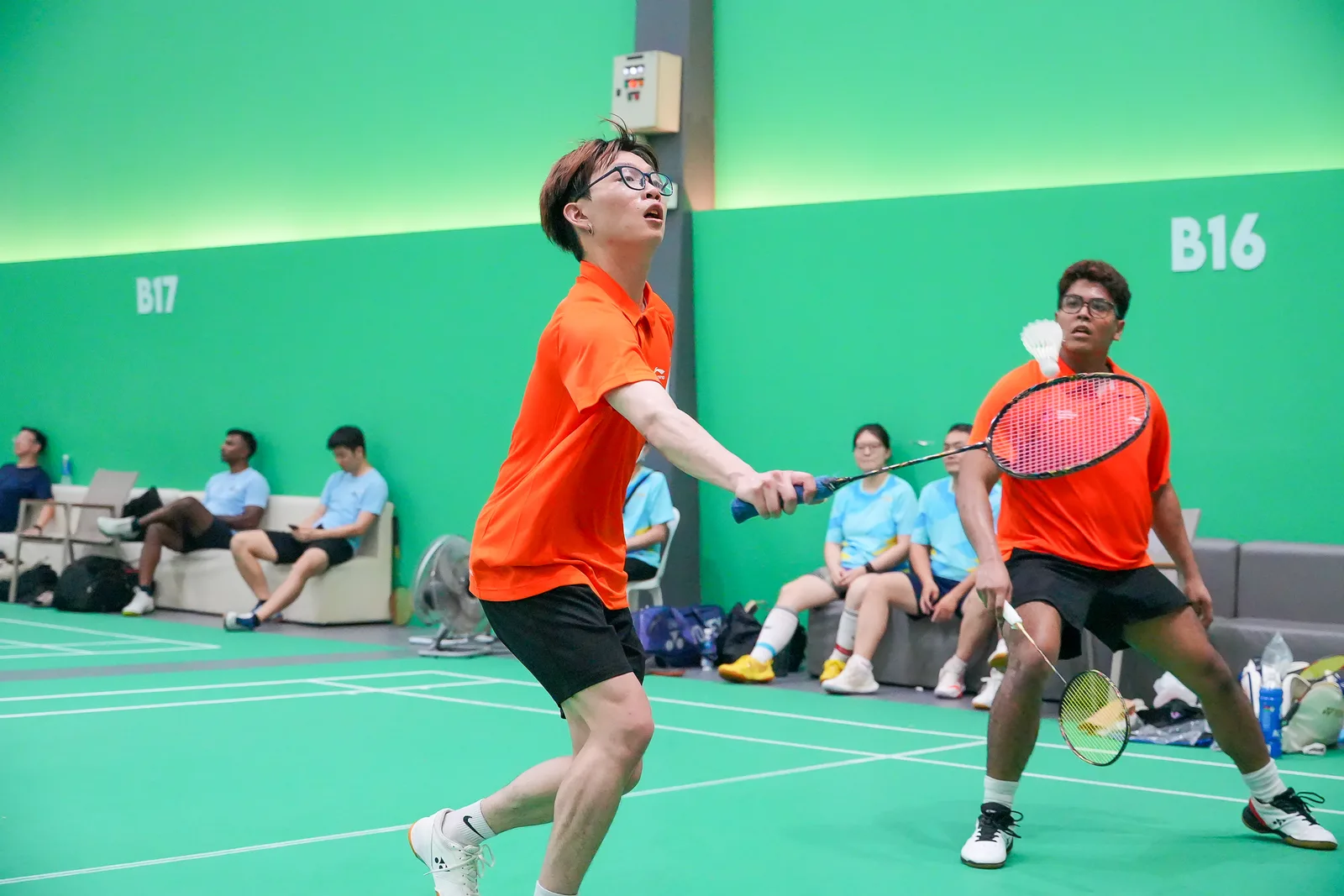 Badminton doubles at moment of contact, orange kits popping against green court — decent action timing, background a little busy with seated