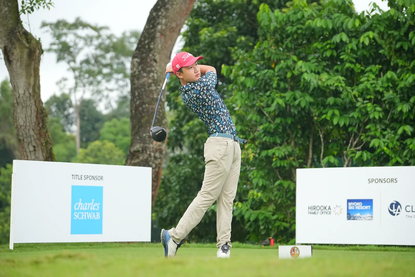 Golf tee shot at full swing finish with title-sponsor boards clean in frame — tidy corporate-sports coverage with sponsor visibility, good t