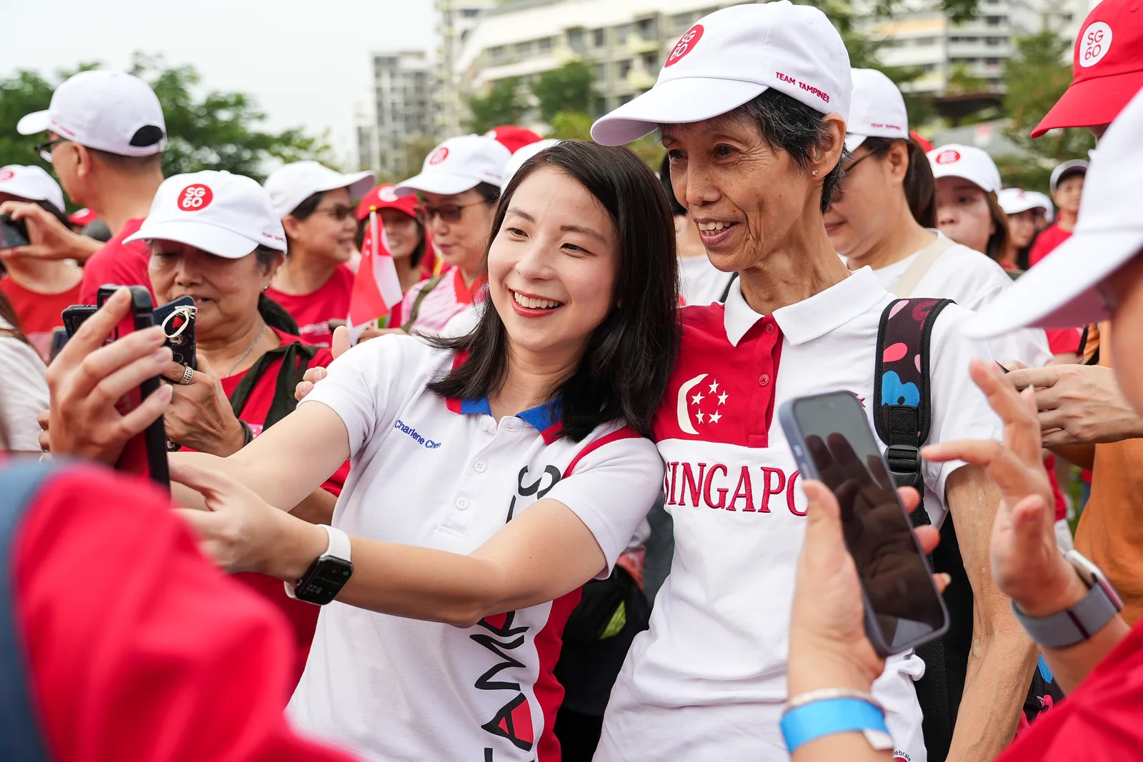Selfie moment between young volunteer and elderly participant at a red-and-white national event — warm, layered, genuinely candid crowd ener