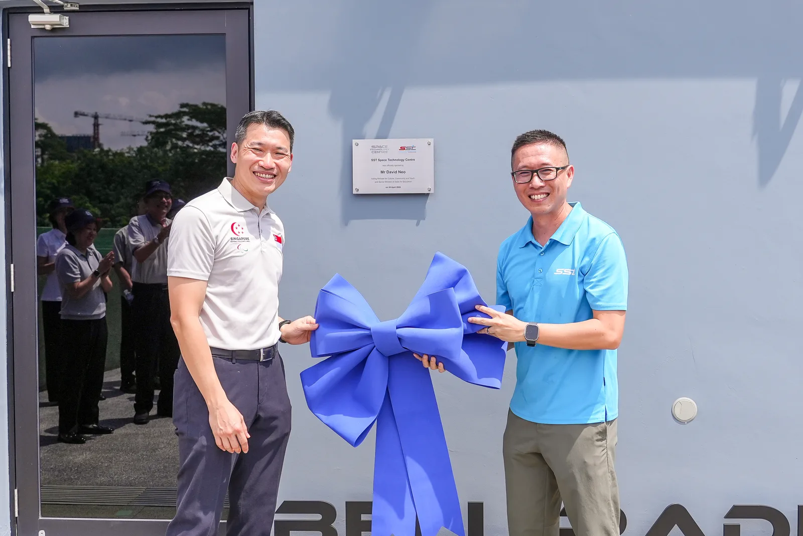 Plaque unveiling with oversized blue bow, both principals engaged and clean symmetry — standard but well-executed corporate opening record
