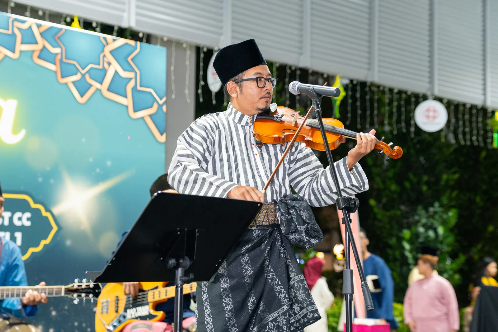 Violinist in traditional Malay dress on stage — nice performance moment, sharp and well-exposed, but community-club setting and busy edges k