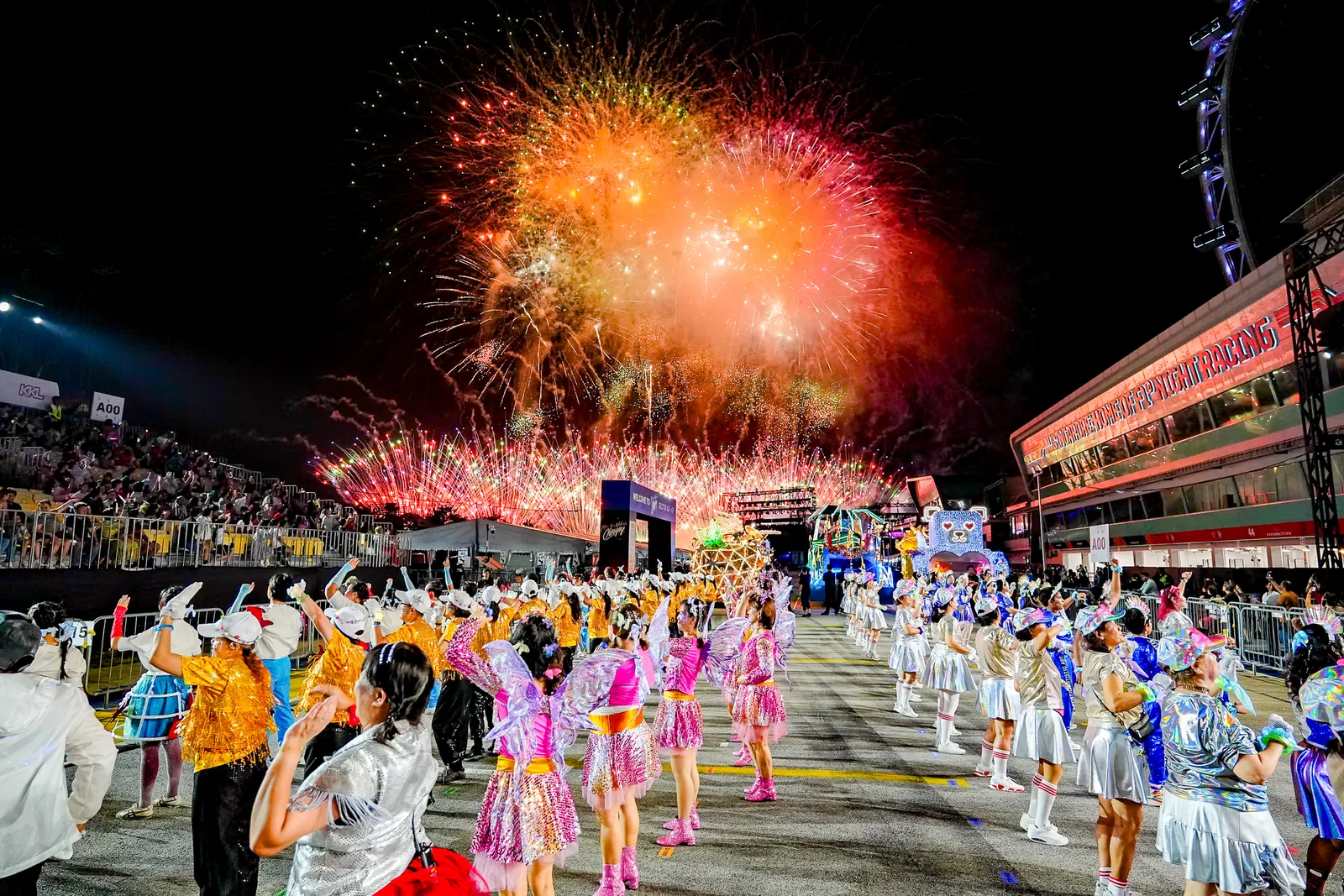 Spectacular night frame — fireworks bursting over a full parade ground with costumed performers and grandstand below