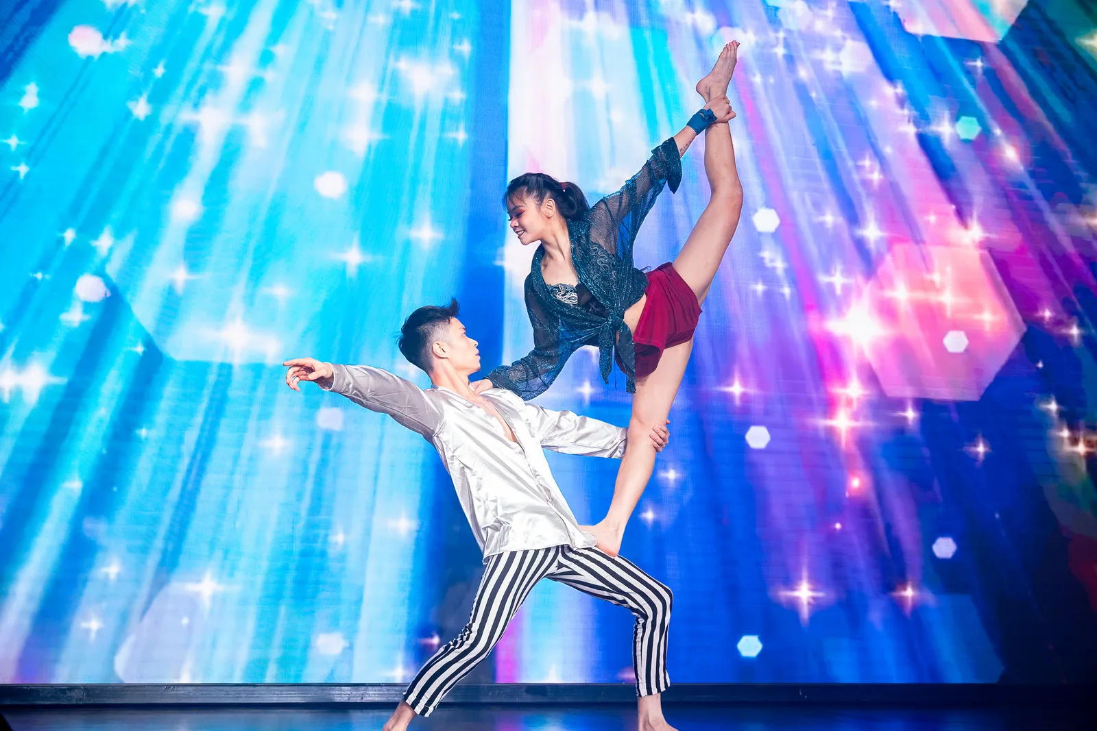 Acrobatic dance duo mid-pose against a blue/purple LED starburst — dramatic stage light, clean silhouette shapes, peak-action timing