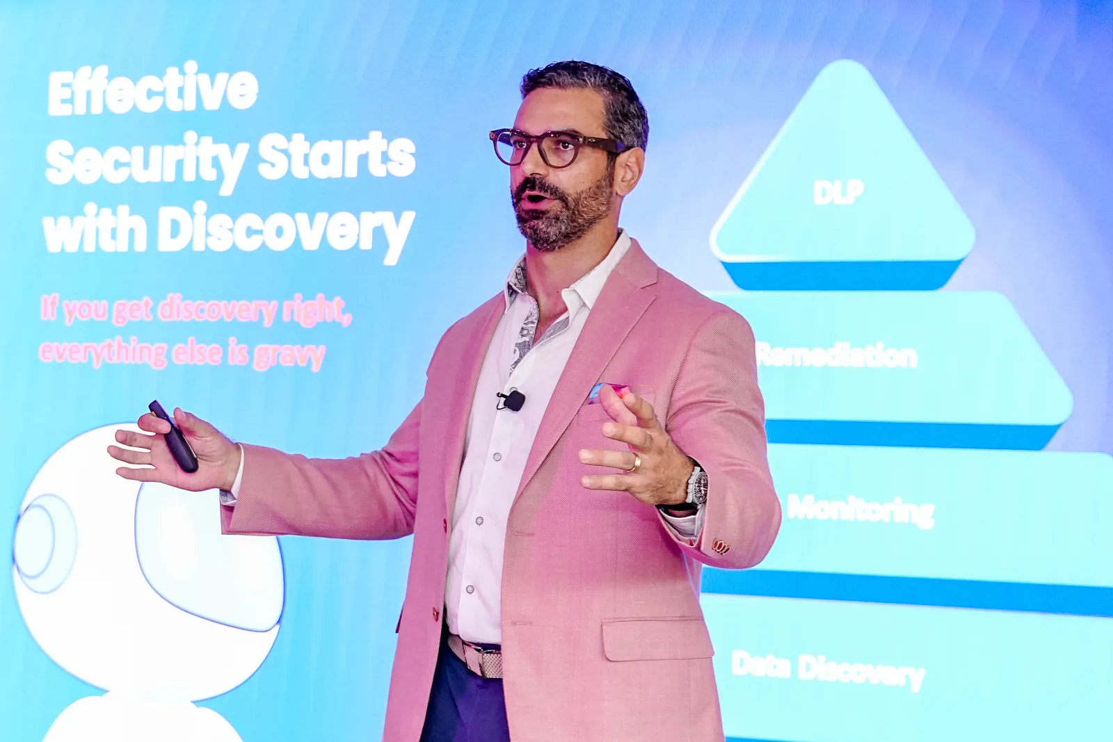 Clean keynote capture — speaker in pink blazer mid-gesture against a saturated blue LED screen, readable slide title, nice colour contrast a