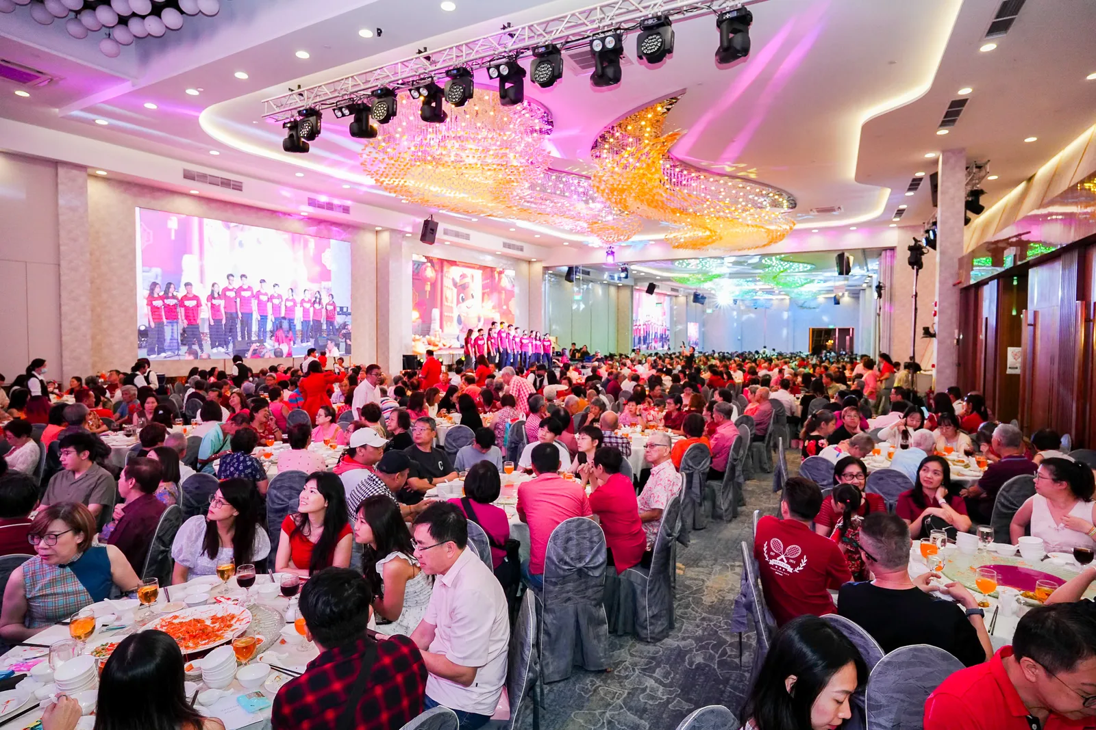 Wide ballroom establishing shot — ornate chandelier venue, pink/purple stage wash, packed tables, performers on stage