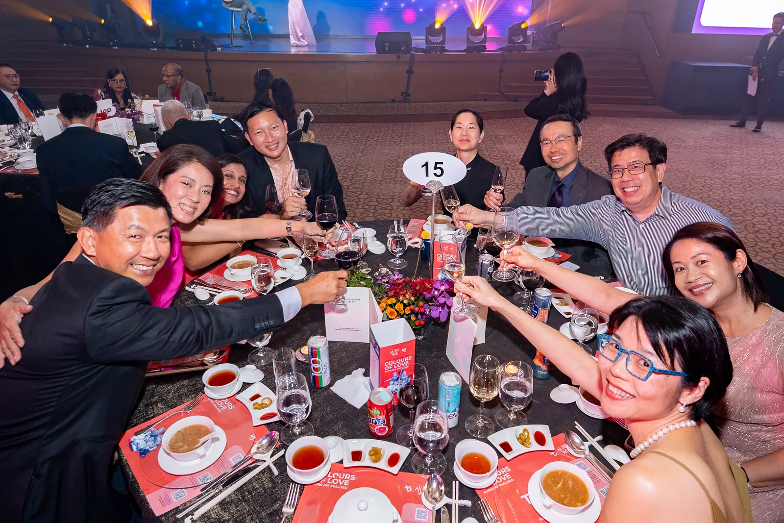 High-energy gala table toast — genuine laughter, glasses raised, dinner-event content