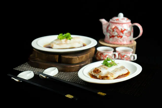 Two plates of chee cheong fun garnished with coriander, arranged on a black background with a red-and-white dragon-pattern teapot, matching cups, chopsticks and spoons, lit low-key against a black background